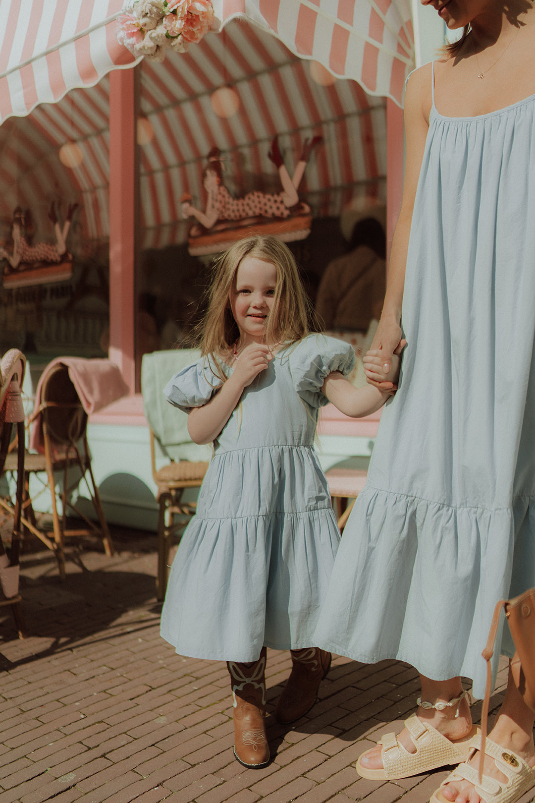 Little girl and mother wearing matching light blue dresses while walking together outside a pastel café in a soft summer setting