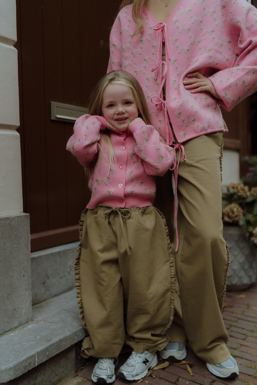 Young girl and adult wearing matching pink floral cardigans and beige pants outdoors.