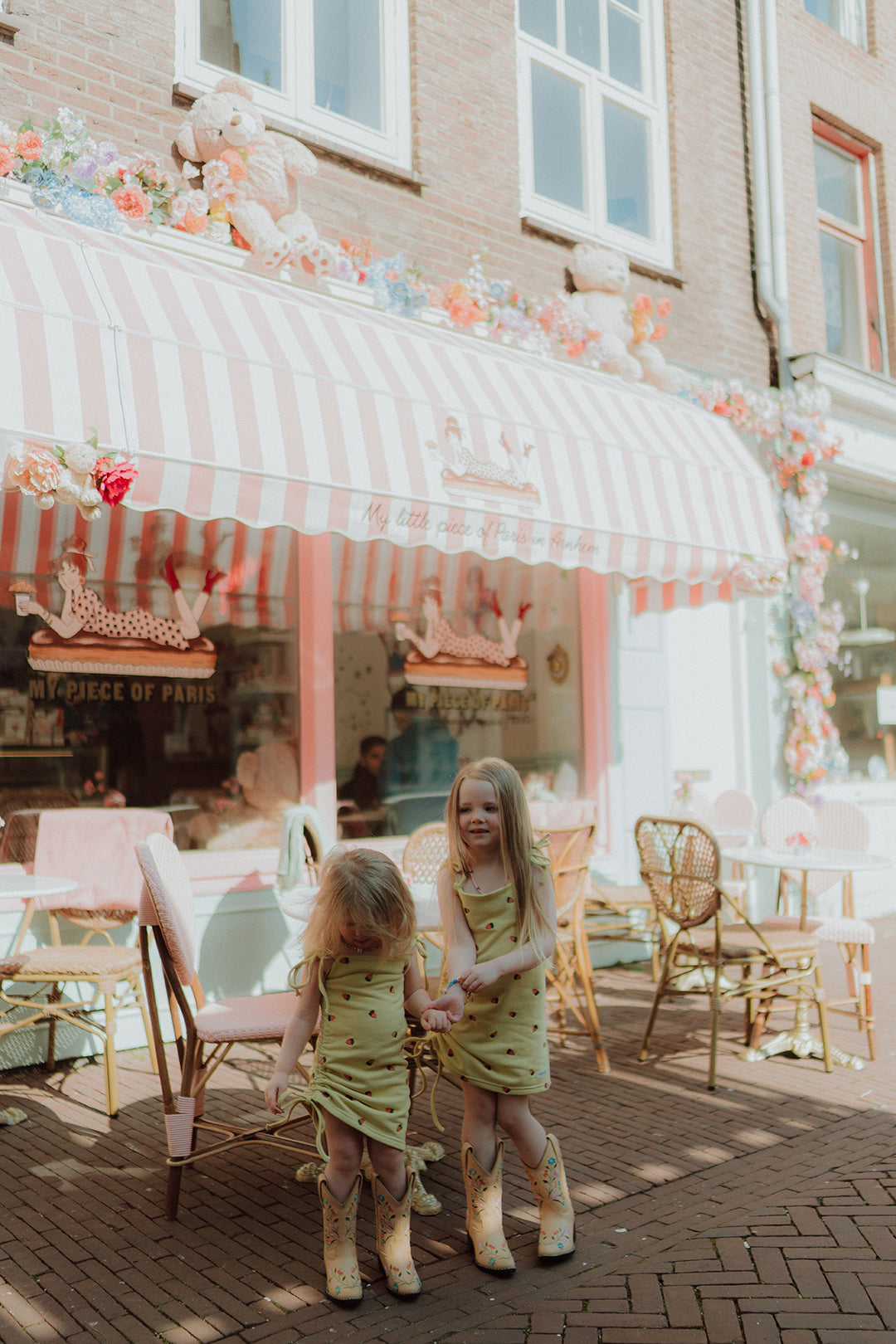 Two little girls wearing matching yellow dresses and cowboy boots outside a pastel café in trendy sisters outfits