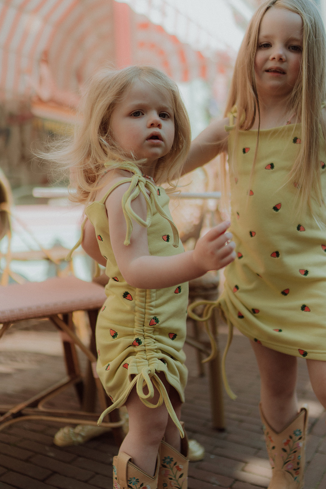 Two little girls wearing matching yellow strawberry print dresses and cowboy boots in a dreamy pastel café setting