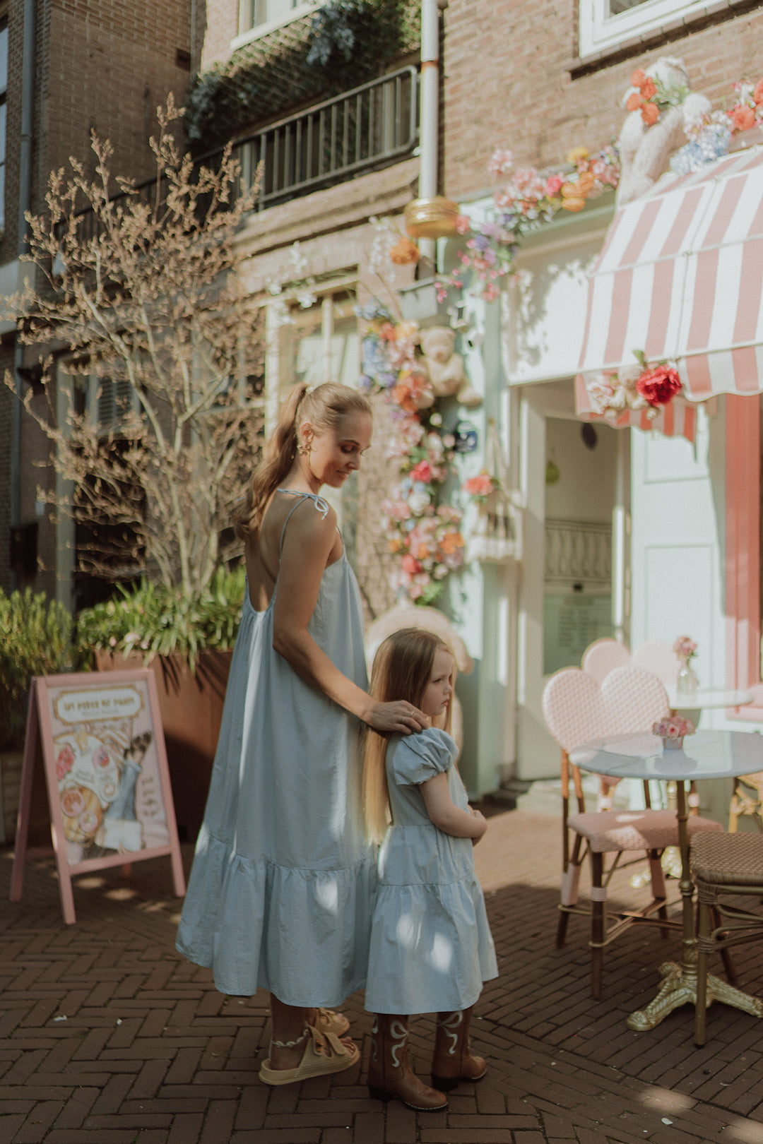 Mother and daughter wearing matching light blue dresses outside a pastel café with floral decorations and a soft dreamy summer aesthetic