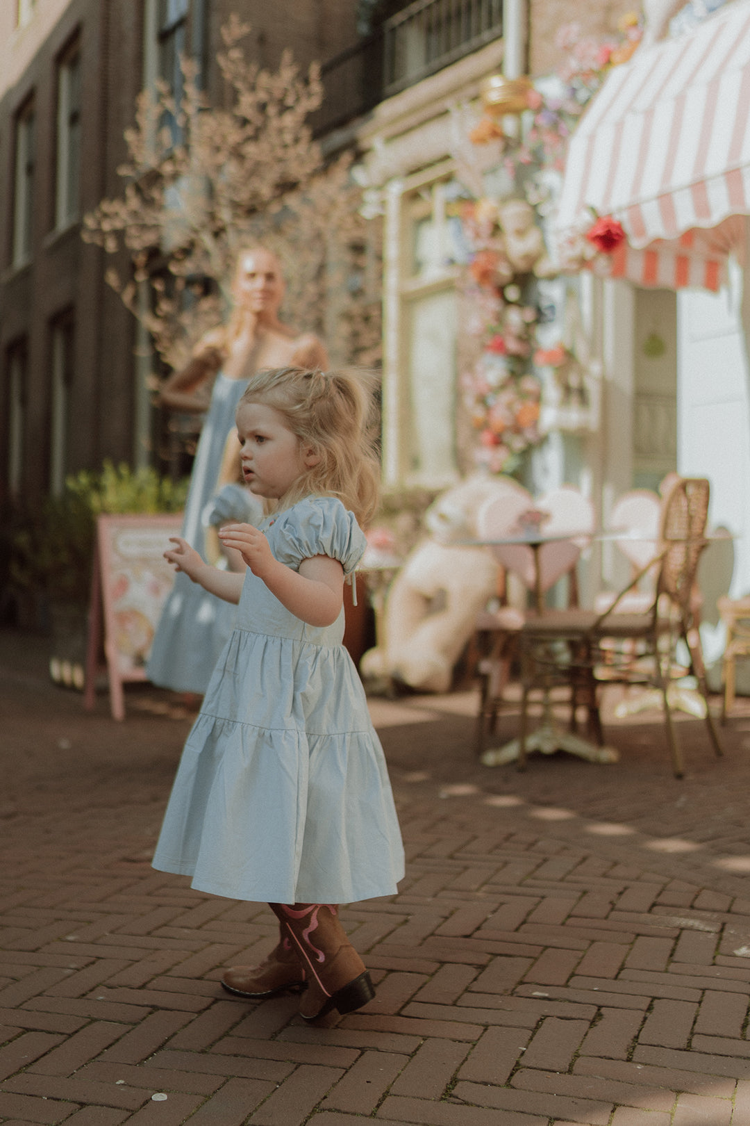Toddler girl in a light blue dress and western boots walking through a charming pastel café street with soft sunlight and dreamy atmosphere.