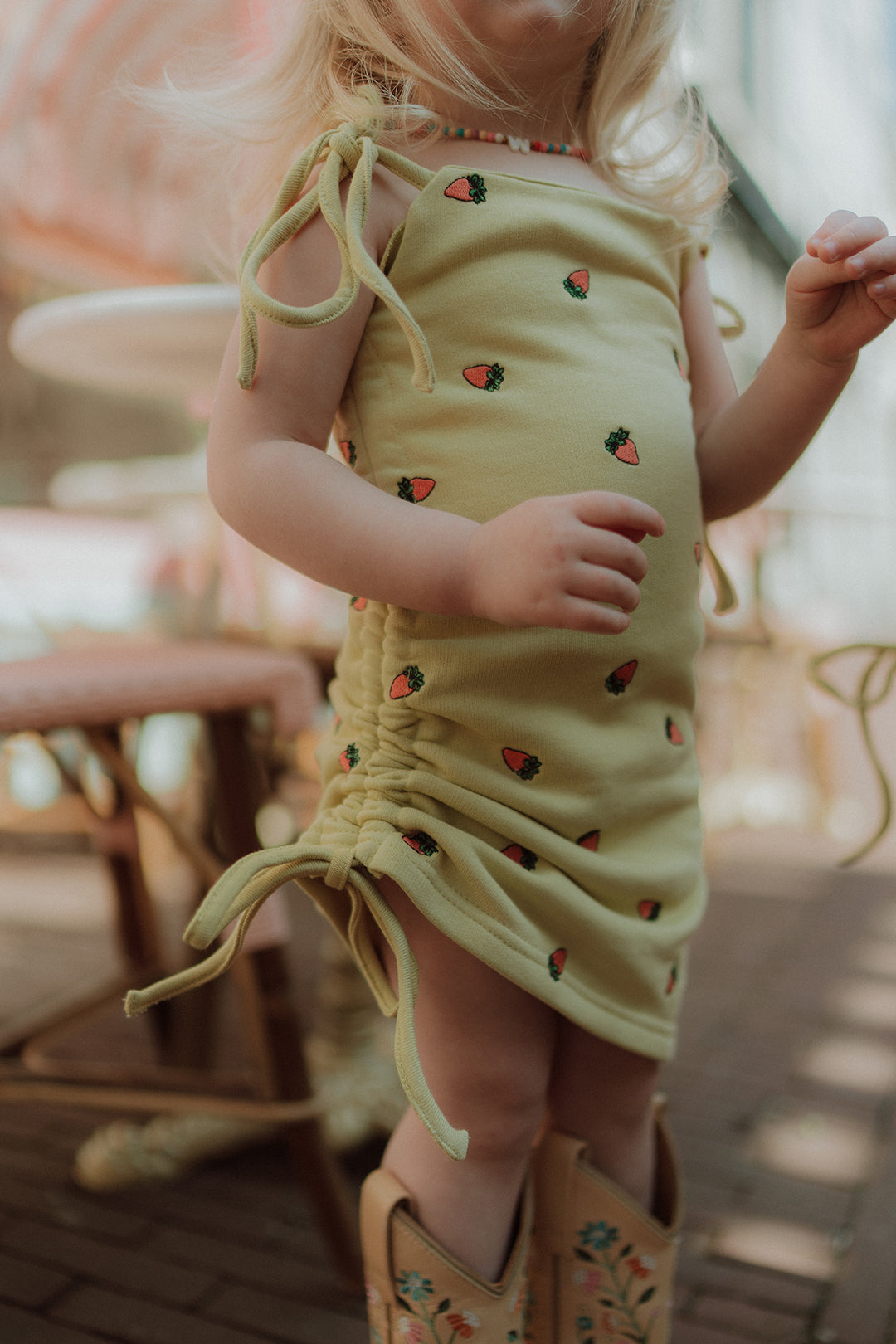 Little girl wearing a yellow strawberry print summer dress with cowboy boots in a dreamy pastel café setting