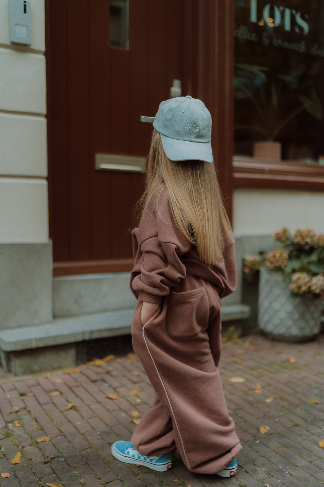 Girl in a mauve oversized tracksuit and blue cap walking outdoors viewed from the back.