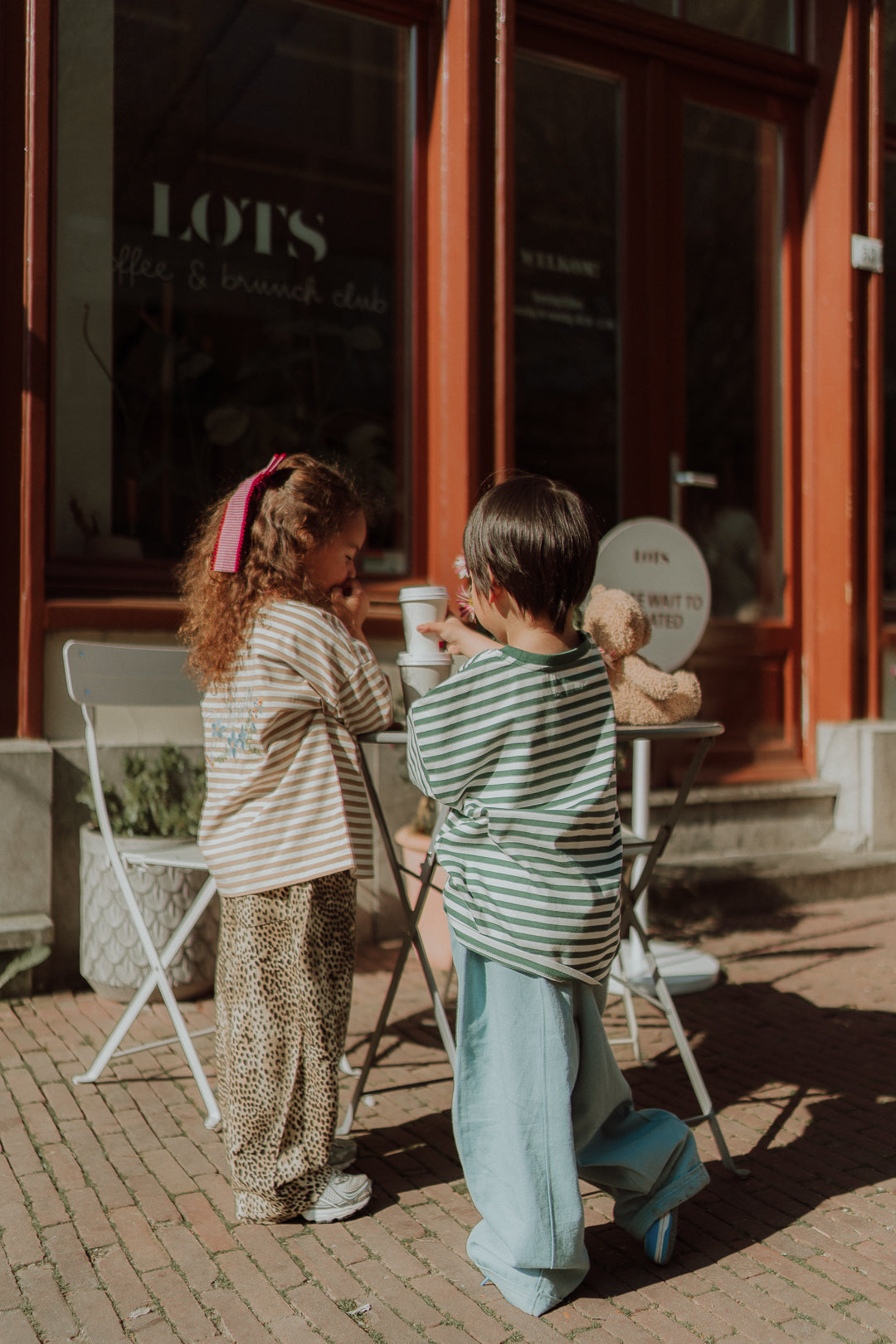 Two children wearing striped oversized shirts and wide leg pants outside a cafe