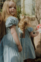 Two little girls wearing matching light blue puff sleeve dresses in a dreamy outdoor summer setting with soft natural light.