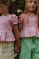 Two girls wearing matching pink smocked tops and patterned pants while holding hands outdoors.