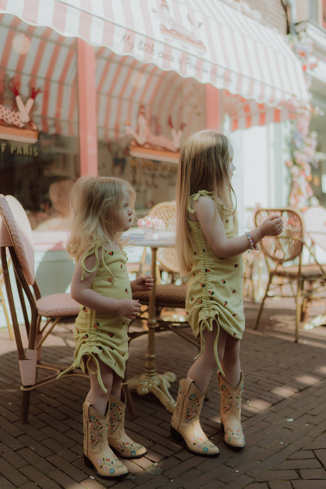 Two little girls wearing matching yellow summer dresses and cowboy boots outside a pastel café with a soft vintage aesthetic