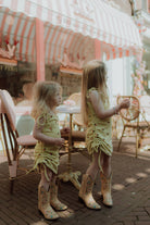 Two little girls wearing matching yellow summer dresses and cowboy boots outside a pastel café with a soft vintage aesthetic