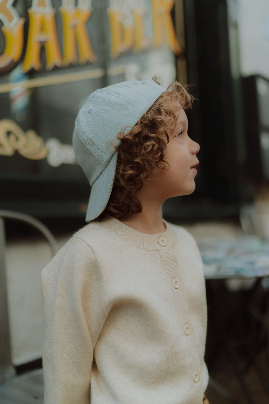 Young child wearing a cream cardigan and light blue cap looking sideways outdoors.
