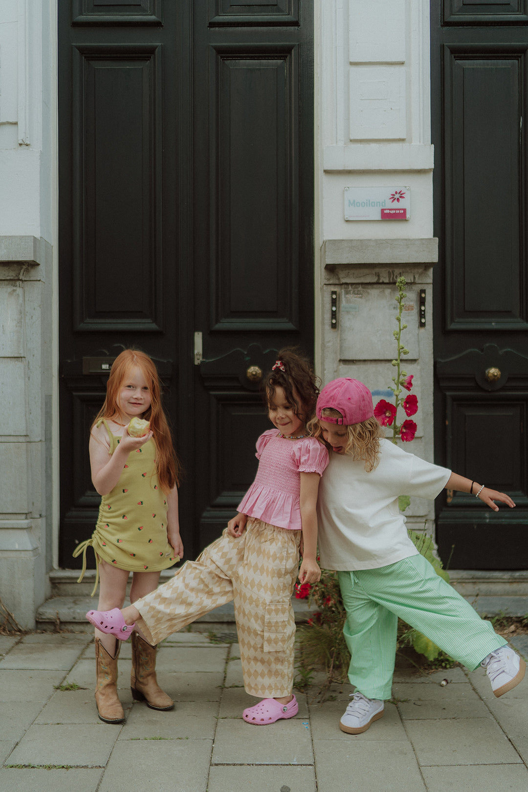 Three kids wearing colorful summer outfits standing and playing in front of black doors outdoors.