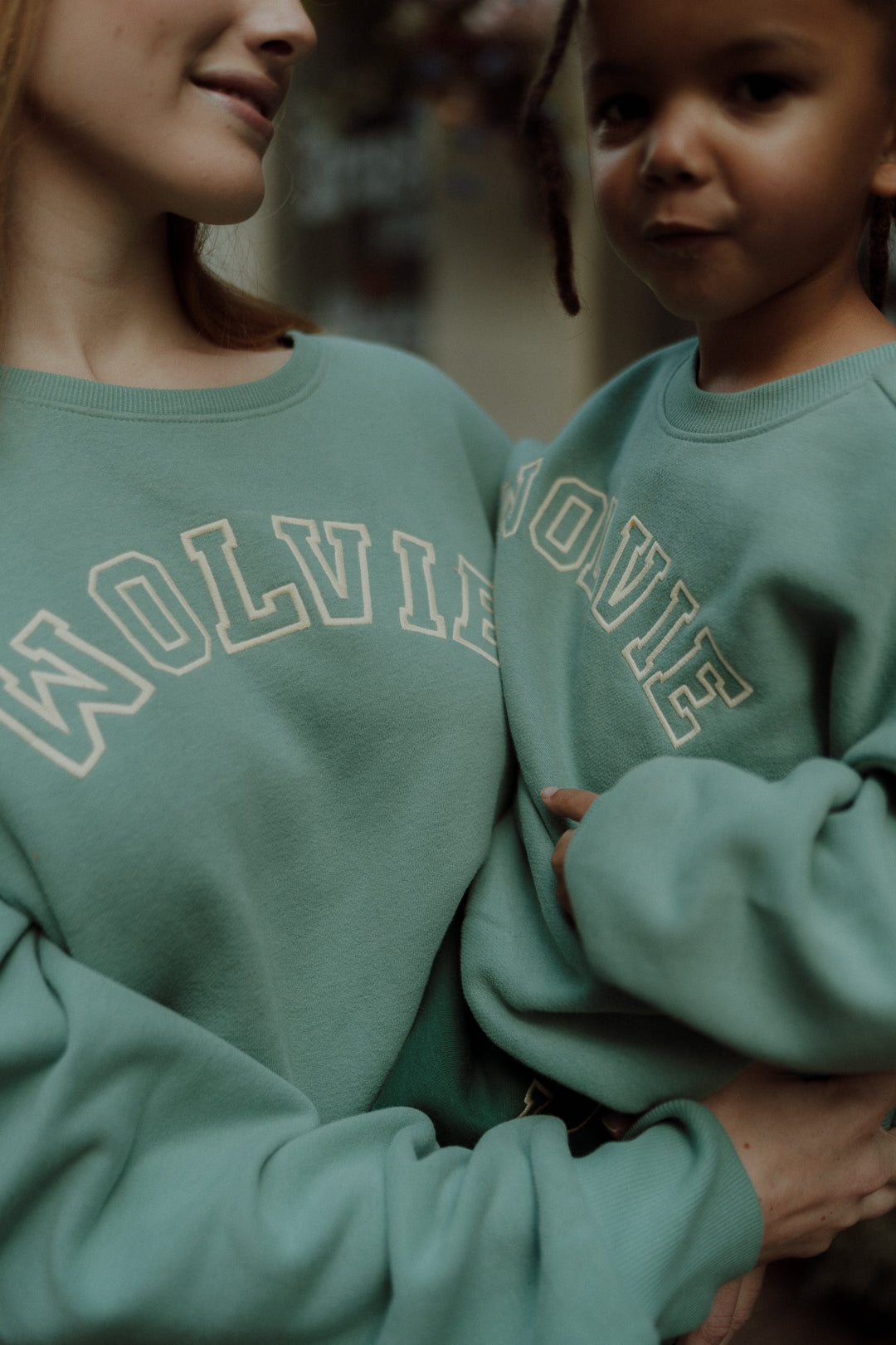 Close-up of an adult and child wearing matching sage green sweatshirts outdoors.
