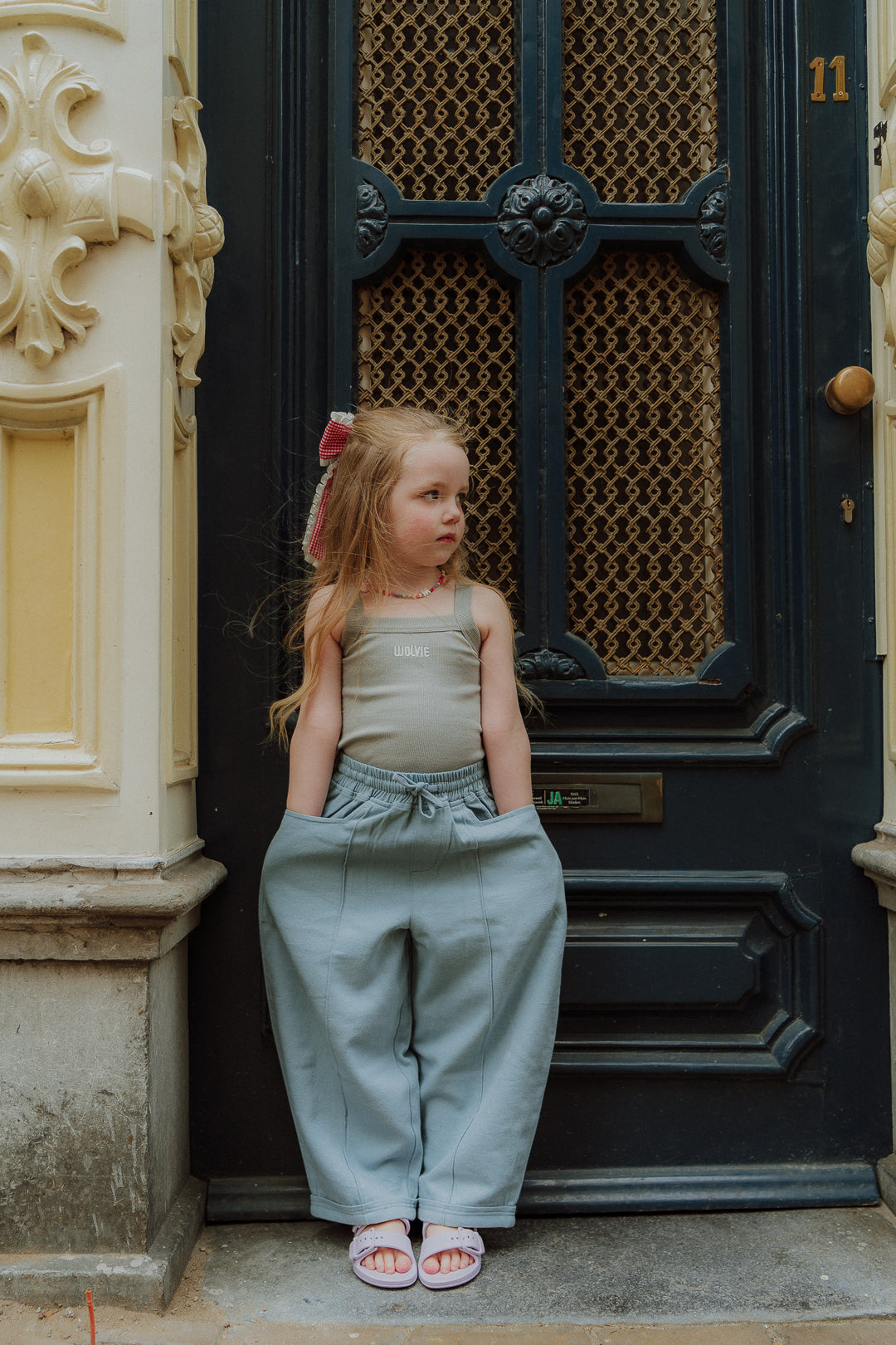 Toddler wearing matching mom and child outfit with green pants and sleeveless top standing in front of a dark door.
