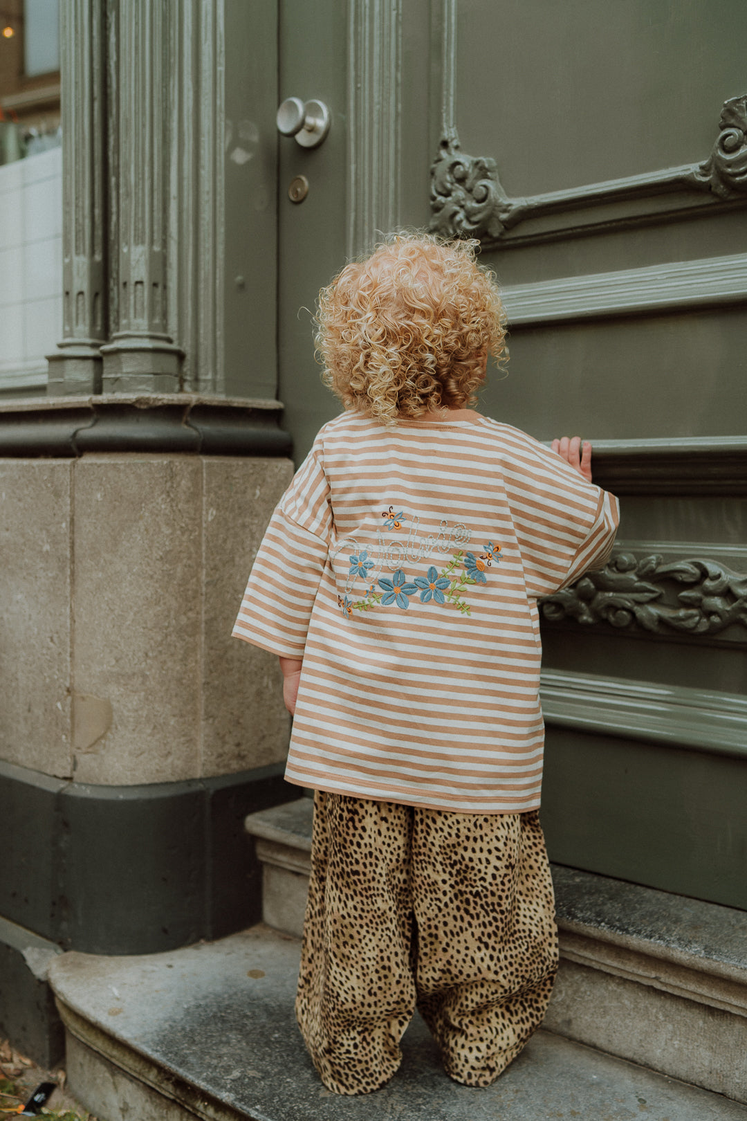 Toddler wearing matching mom and child outfit with striped embroidered shirt and leopard print pants seen from the back.