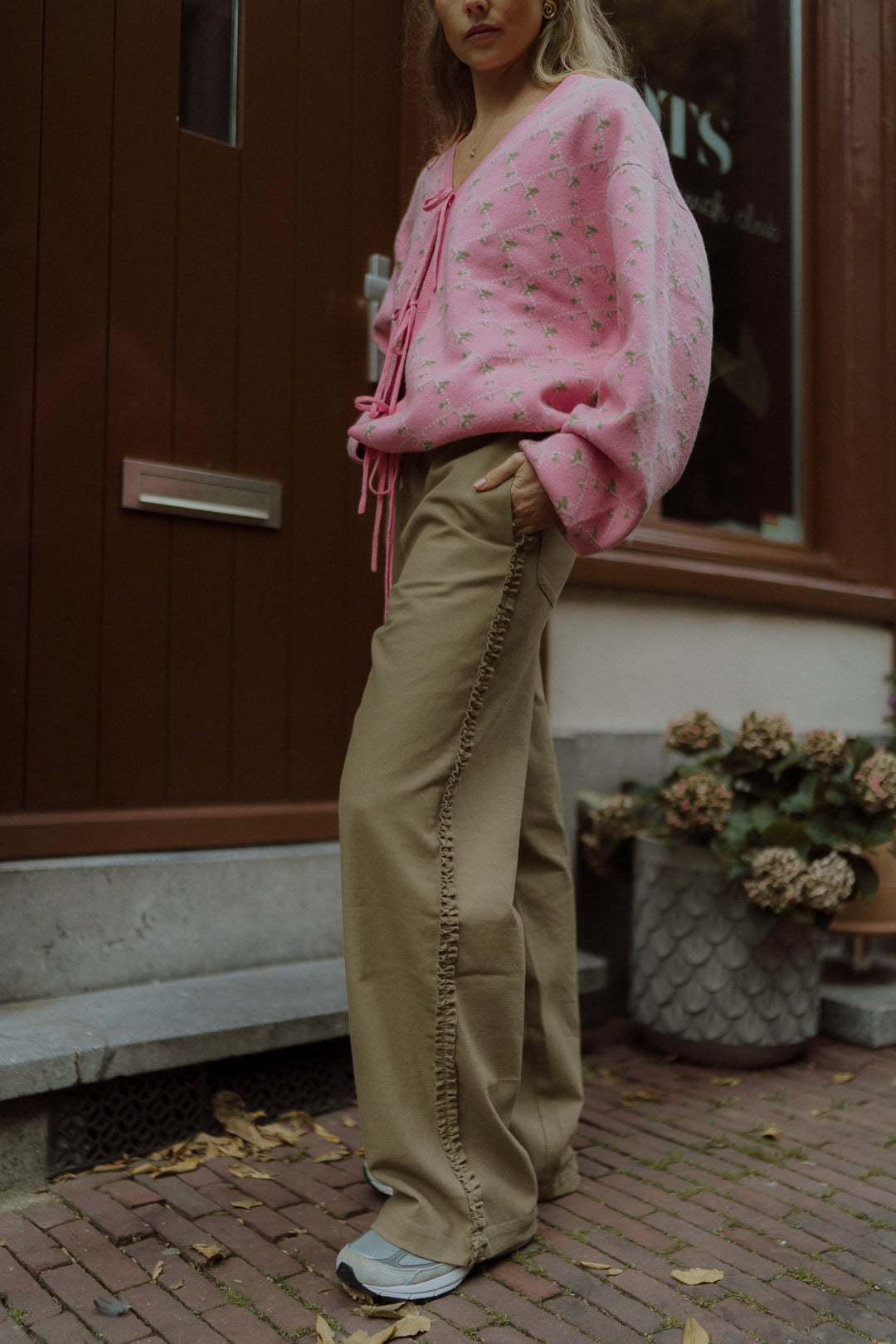 Side view of a woman wearing a pink floral cardigan with beige ruffle-detail pants outdoors.