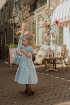 Toddler girl in a light blue dress and western boots walking through a charming pastel café street with soft sunlight and dreamy atmosphere.