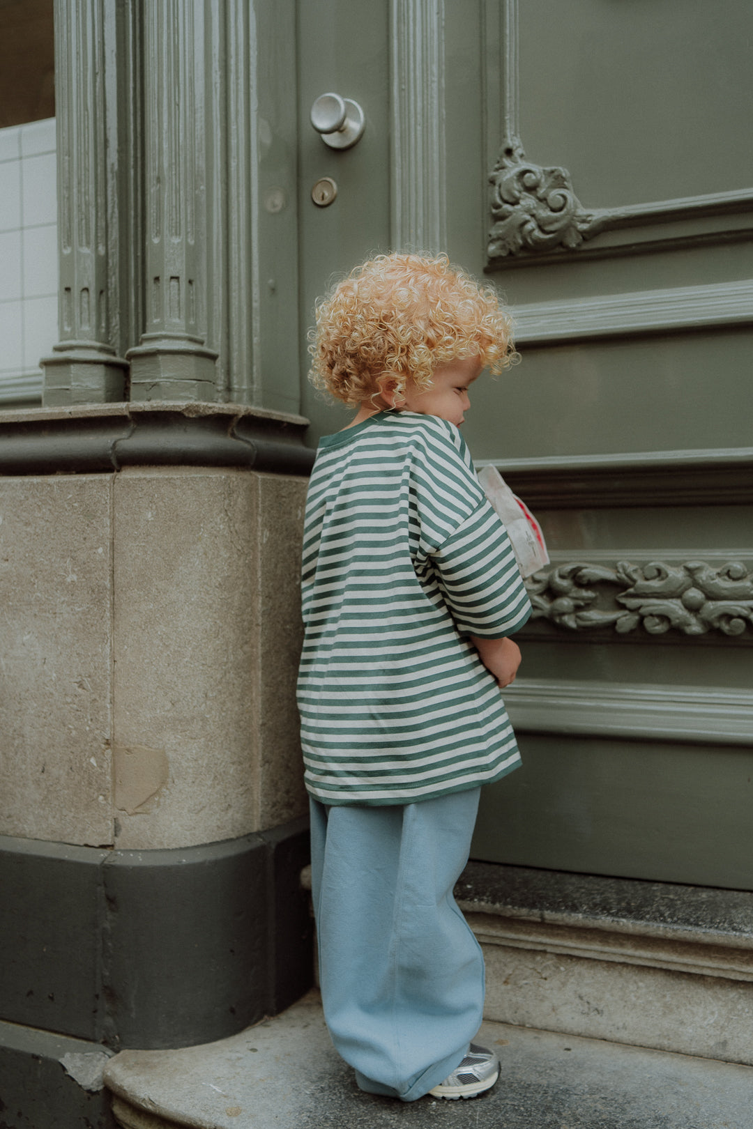 Toddler wearing green striped shirt and loose blue pants standing on a doorstep.