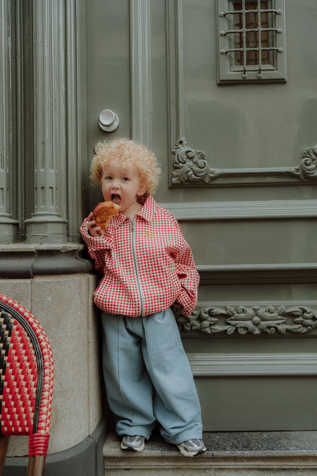 Toddler wearing red gingham jacket and blue pants while eating a pastry